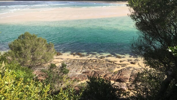 A coastal landscape with clear waters, sandy beach, and rocky outcrops.