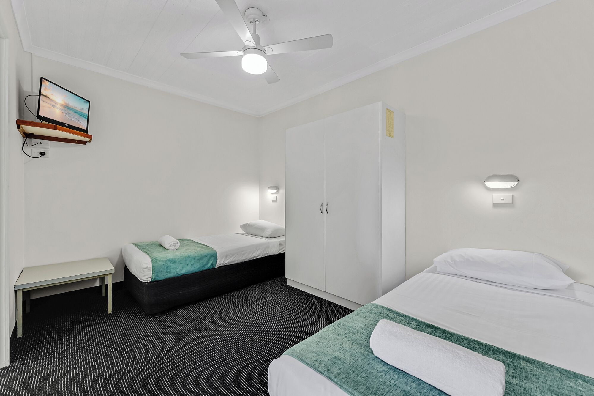 A hotel room with two beds, a desk, a ceiling fan, and a TV mounted on the wall.