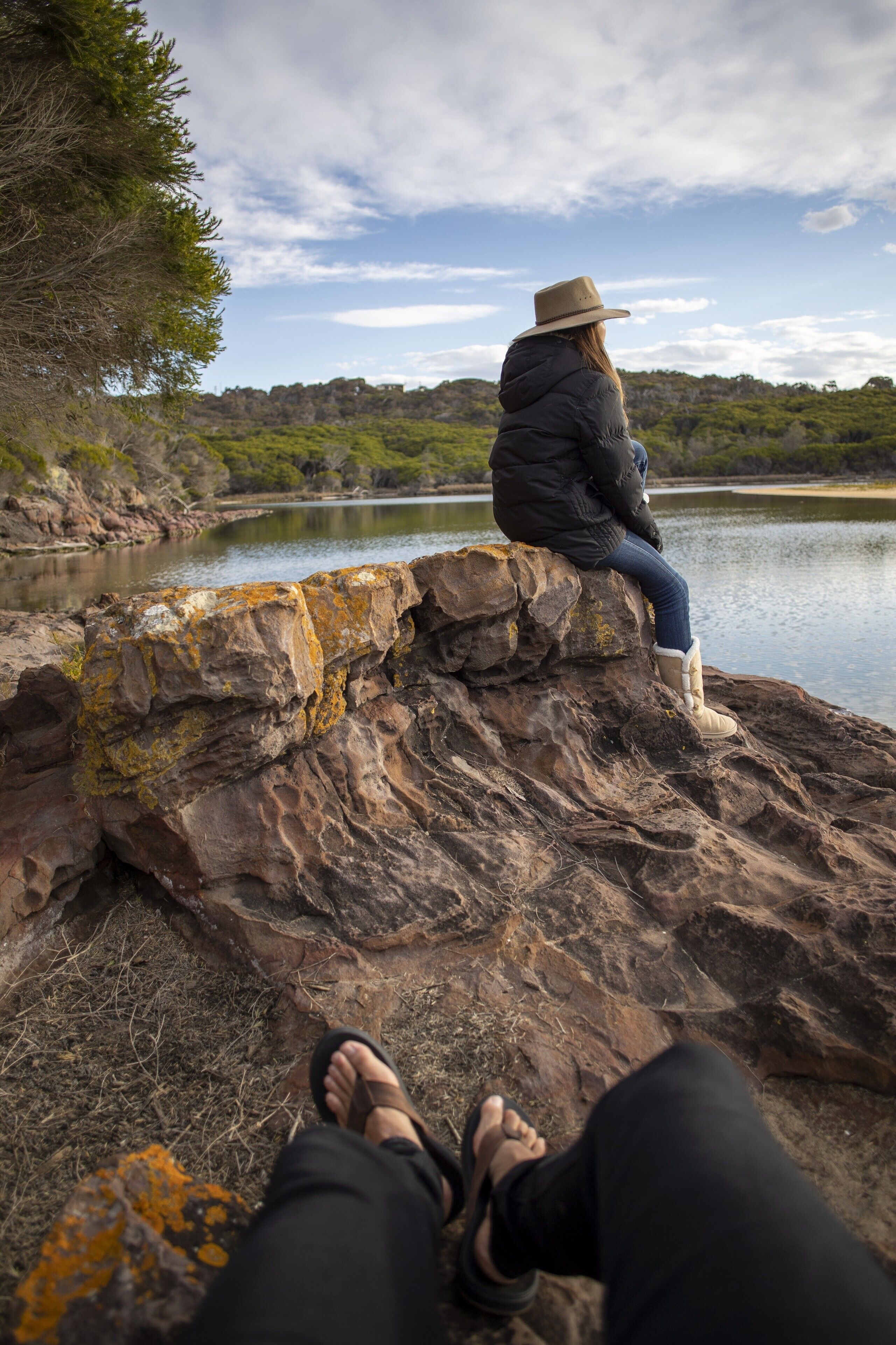 A person sitting on a rocky ledge overlooking a body of water with trees and hills in the background.