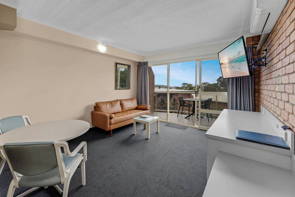 A modern room with a leather sofa, a dining table with chairs, a TV mounted on the wall, and a balcony with a view.