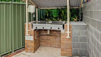 Un barbecue extérieur doté d’un toit en métal et d’une base en briques, entouré d’une clôture verte et de plantes.