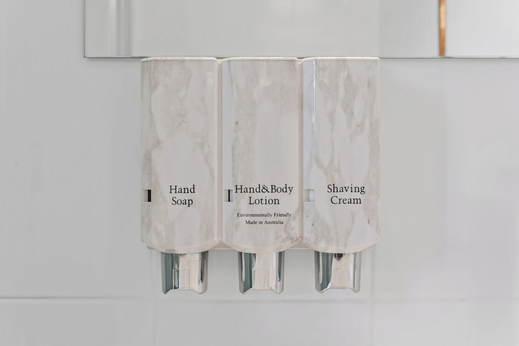 Trois distributeurs de produits d’hygiène, étiquetés « Savon pour les mains », « Lait pour les mains et le corps » et « Crème à raser », sont fixés à un mur blanc.
