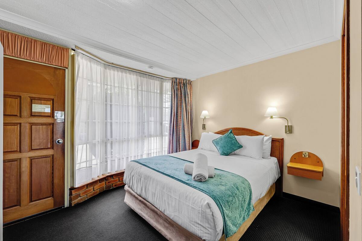 A hotel room with a wooden door, a large bed with a teal bedspread, white bedding, and two wall-mounted lamps.