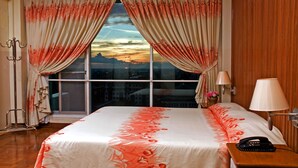 Habitación de hotel con un ventanal grande, cortinas, una cama con ropa de cama floral, una mesita de noche con una lámpara y un teléfono.