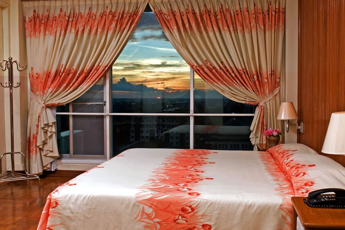 A hotel room with a large window, curtains, a bed with floral bedding, a bedside table with a lamp, and a phone.