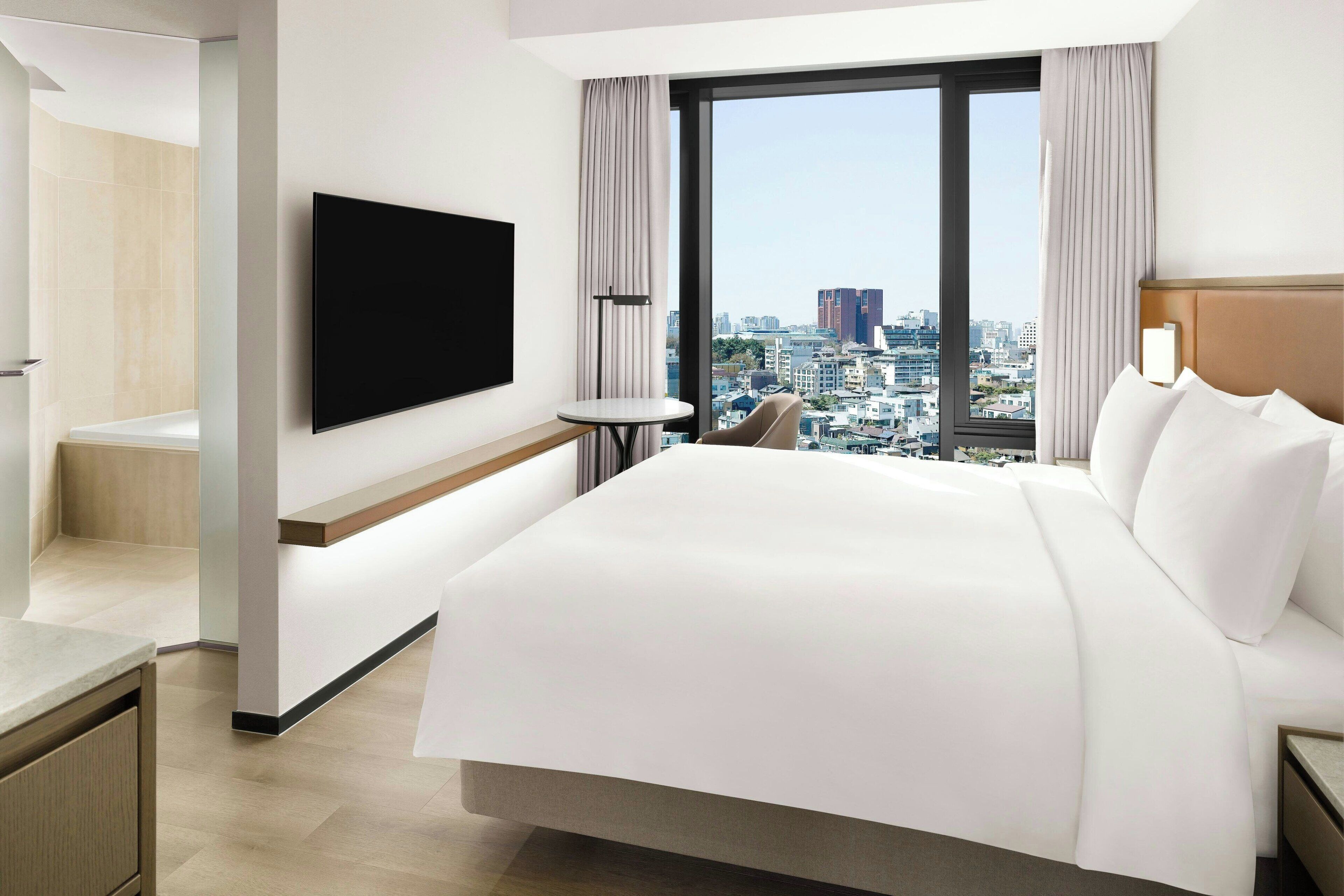 A modern hotel room with a large bed, a flat-screen TV mounted on the wall, a small round table, and a view of the city through large windows.