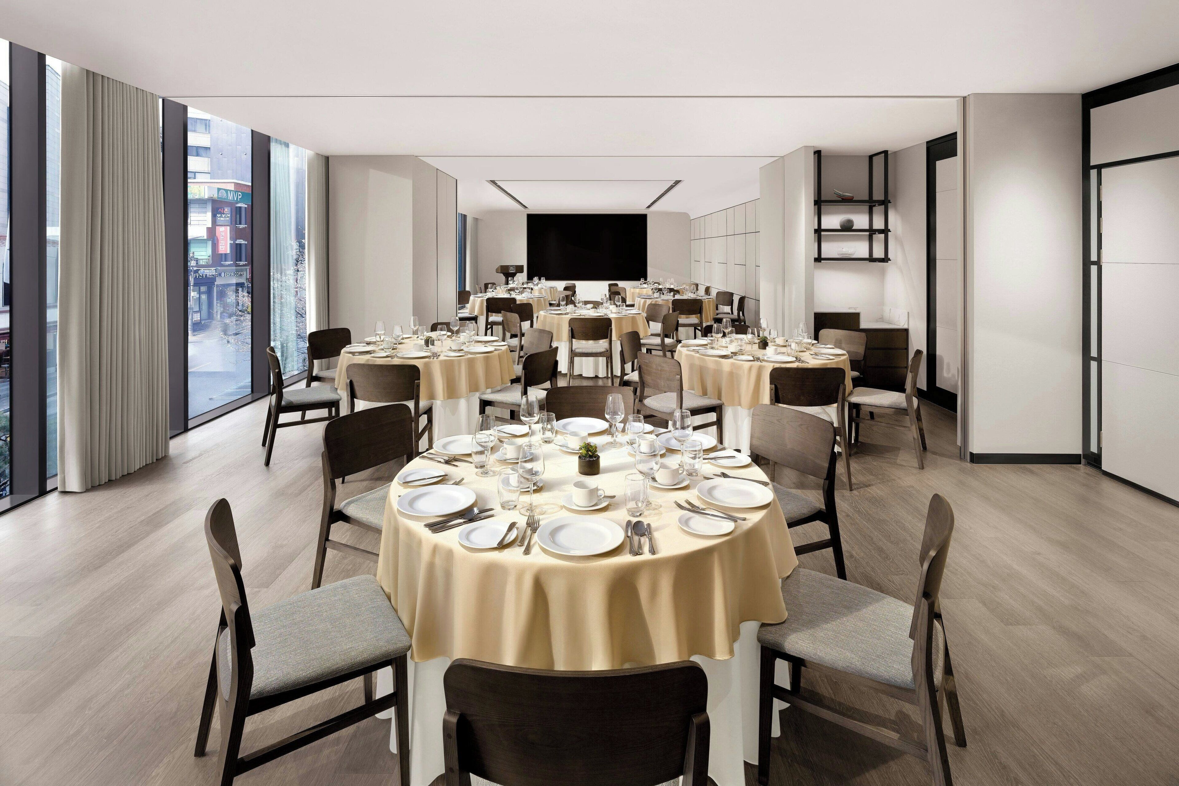 A modern dining area with round tables set for multiple guests, large windows offering a city view, and a flat-screen TV mounted on the wall.