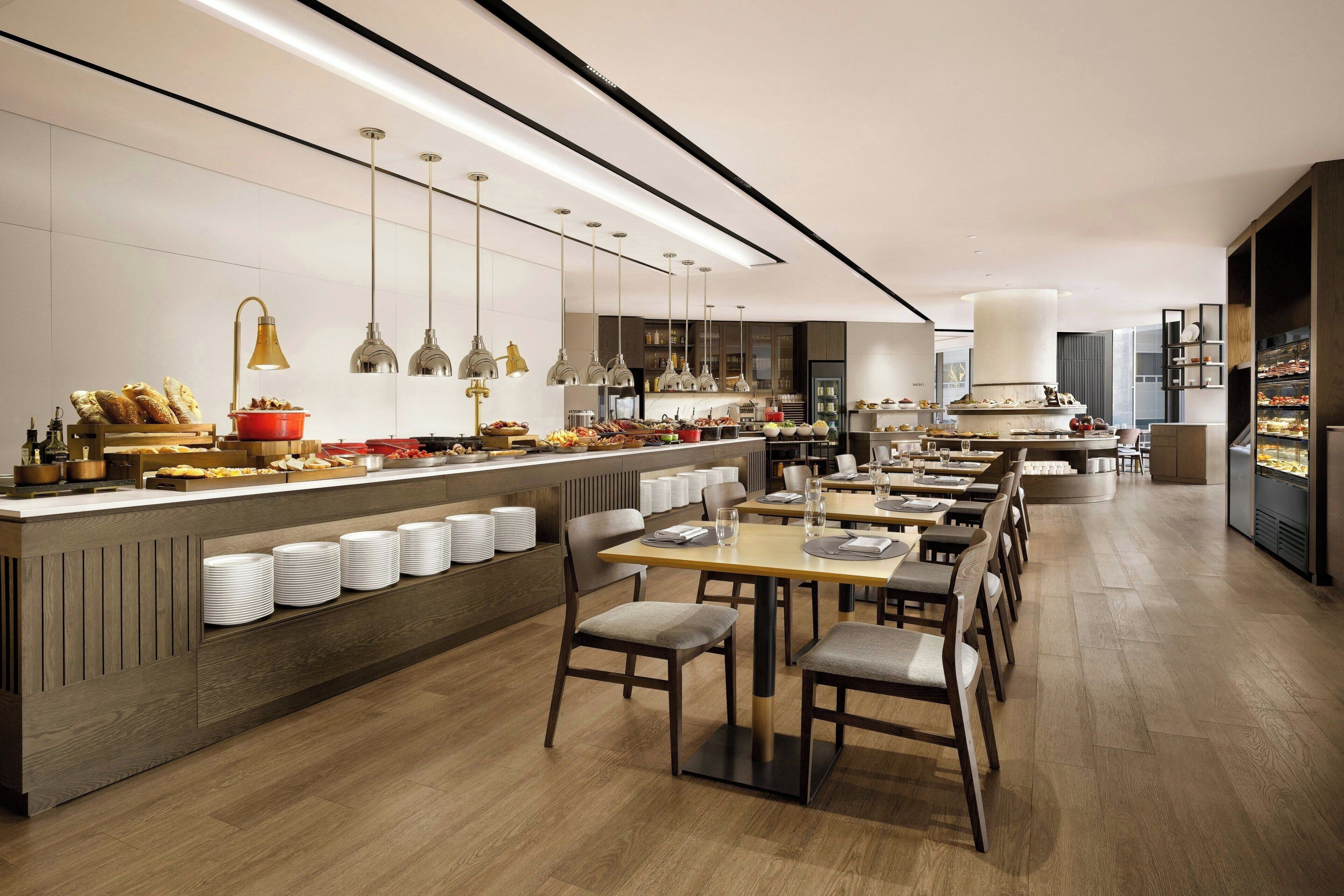 A modern dining area with a long counter, multiple dining tables, and a variety of food items displayed.