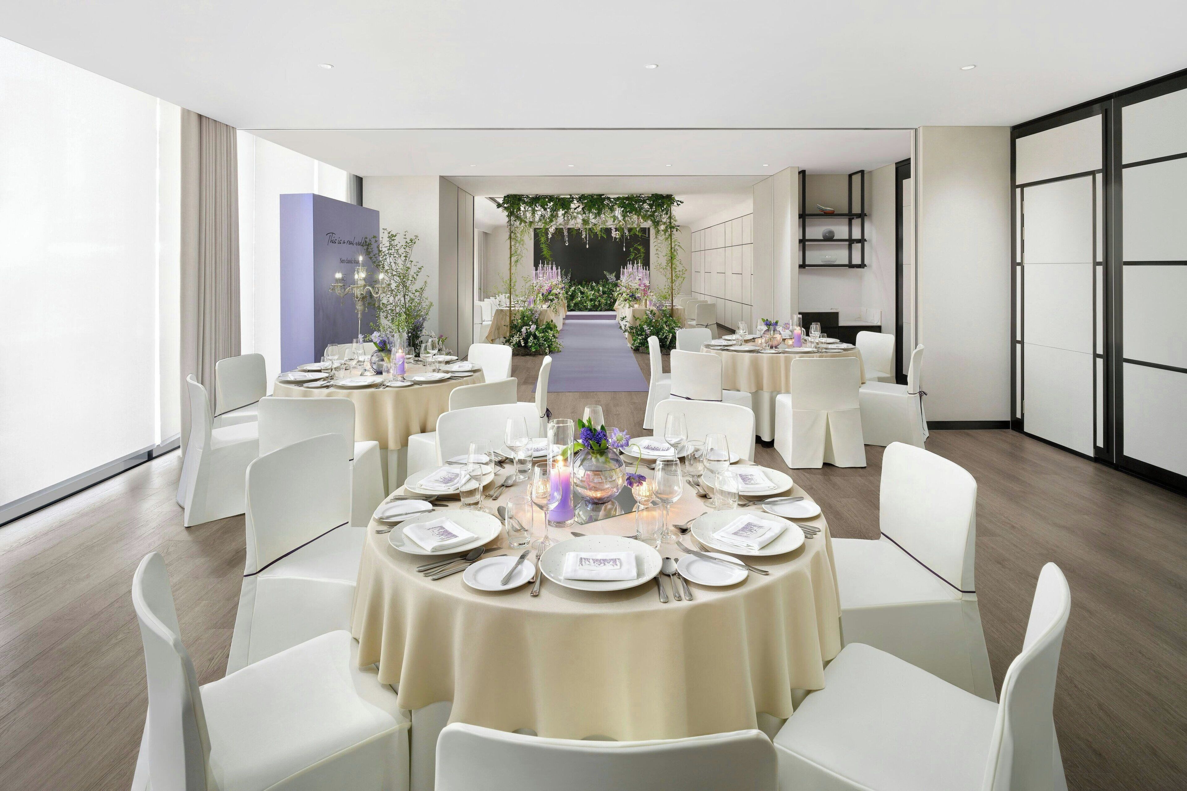A modern dining area with round tables set for a meal, white chairs, and a central aisle leading to a decorative archway.