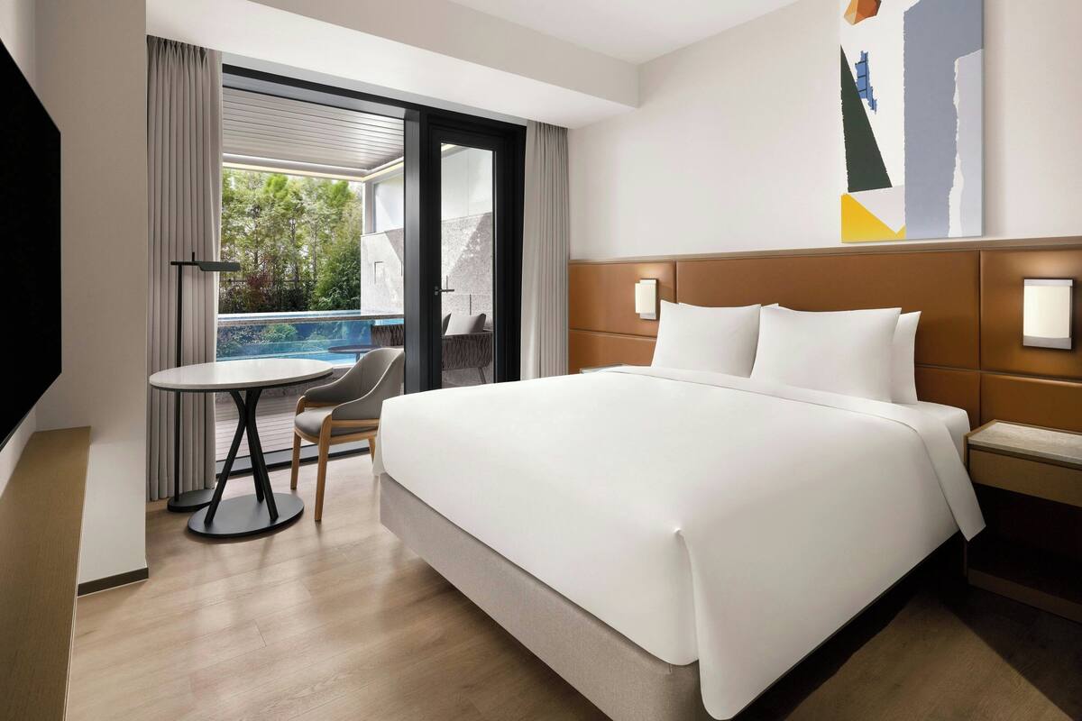 Une chambre d’hôtel avec un grand lit, une petite table, une chaise, une table de chevet et une vue sur une piscine et la verdure environnante.