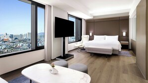 A modern hotel room with a large window offering a city view, a flat-screen TV, a bed, a seating area, and a small table with a vase. - AC Hotel by Marriott Seoul Gangnam (Seoul)