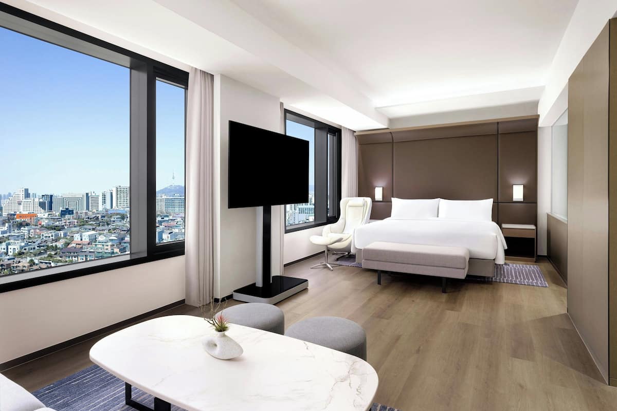 Une chambre d’hôtel moderne dotée d’une grande fenêtre donnant sur la ville, d’un téléviseur à écran plat, d’un lit, d’un coin salon et d’une petite table agrémentée d’un vase.