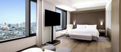 Una habitación de hotel moderna con un ventanal grande que ofrece vistas a la ciudad, un televisor de pantalla plana, una cama, una zona de estar y una pequeña mesa con un jarrón.