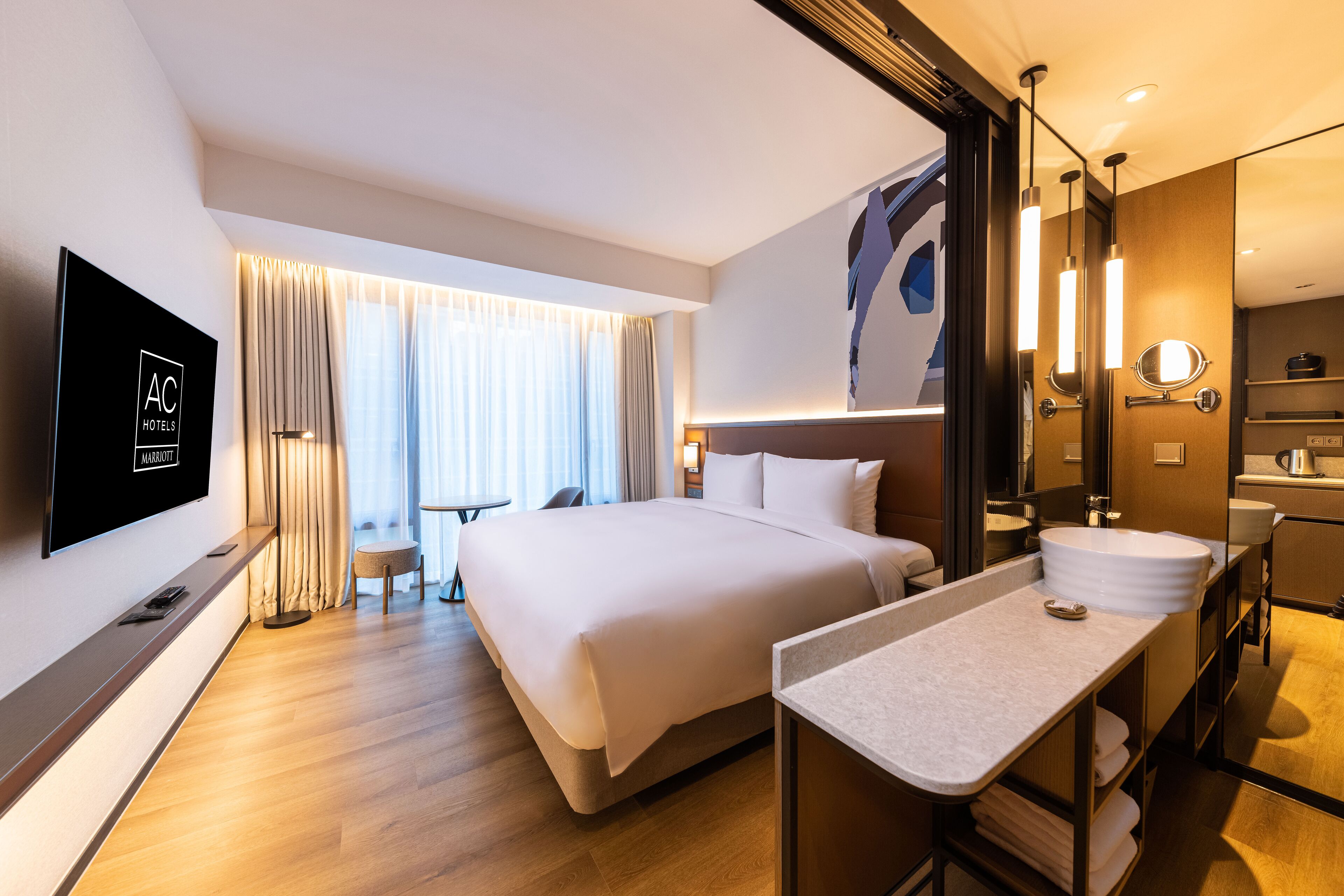 A modern hotel room with a large bed, a flat-screen TV, a bathroom with a sink and mirror, and a window with curtains.