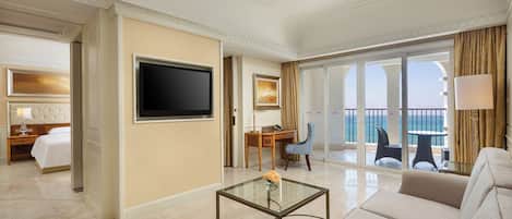 Une chambre d’hôtel dotée d’une grande télévision, d’un canapé, d’une table basse en verre, d’un bureau avec une chaise et d’un balcon offrant une vue sur la mer.