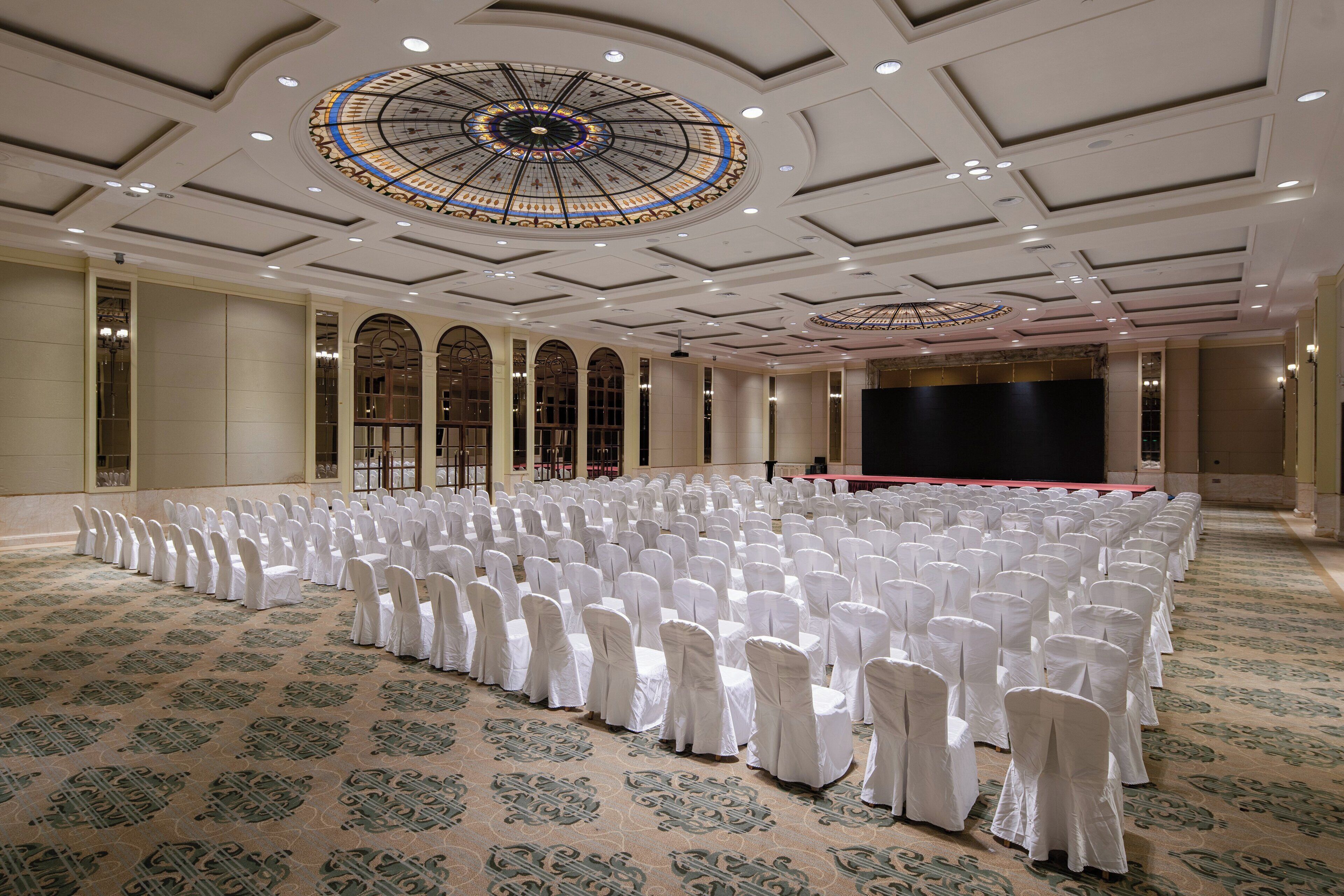 A large hall with rows of white-covered chairs, a stage, and a decorative ceiling.