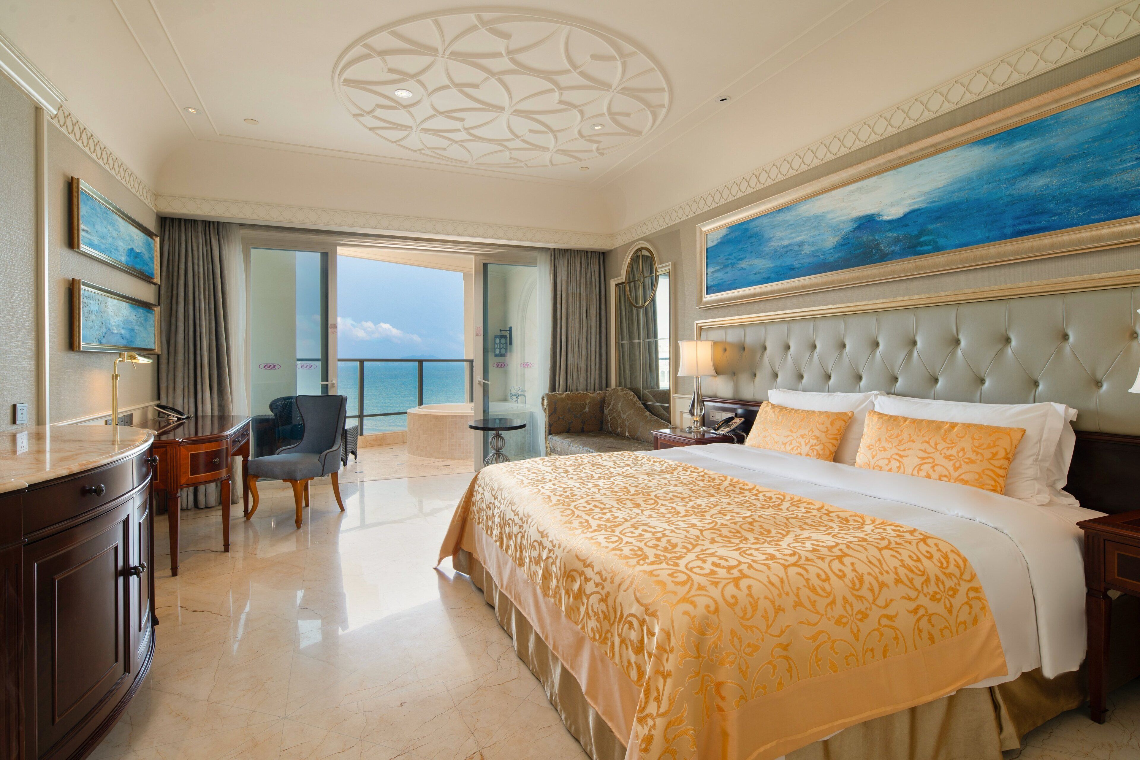A spacious hotel room with a large bed, a sitting area with a chair and a small table, a desk, and a balcony with a view of the sea.