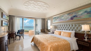 Une chambre d’hôtel spacieuse avec un grand lit, un coin salon avec un fauteuil, un bureau et un balcon offrant une vue sur la mer.