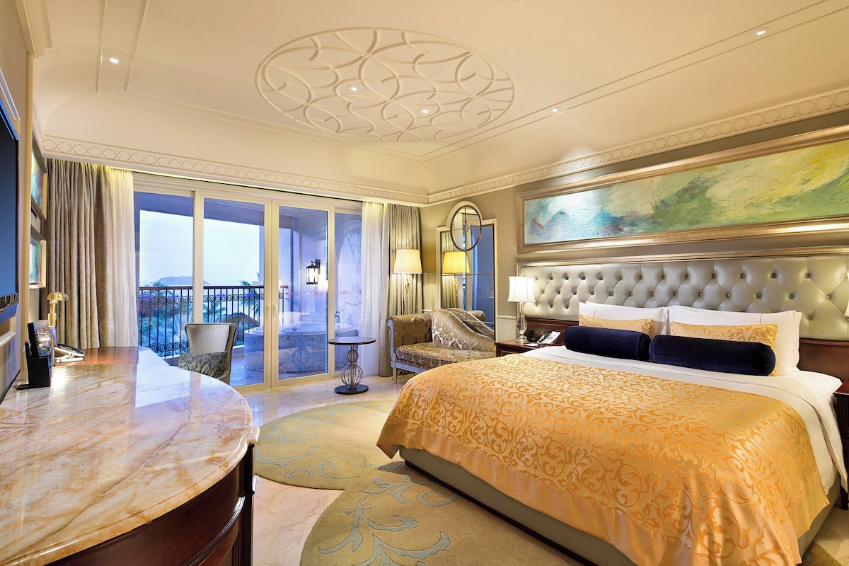 A hotel room with a large bed, a round marble table, a balcony with a view, and a painting on the wall.