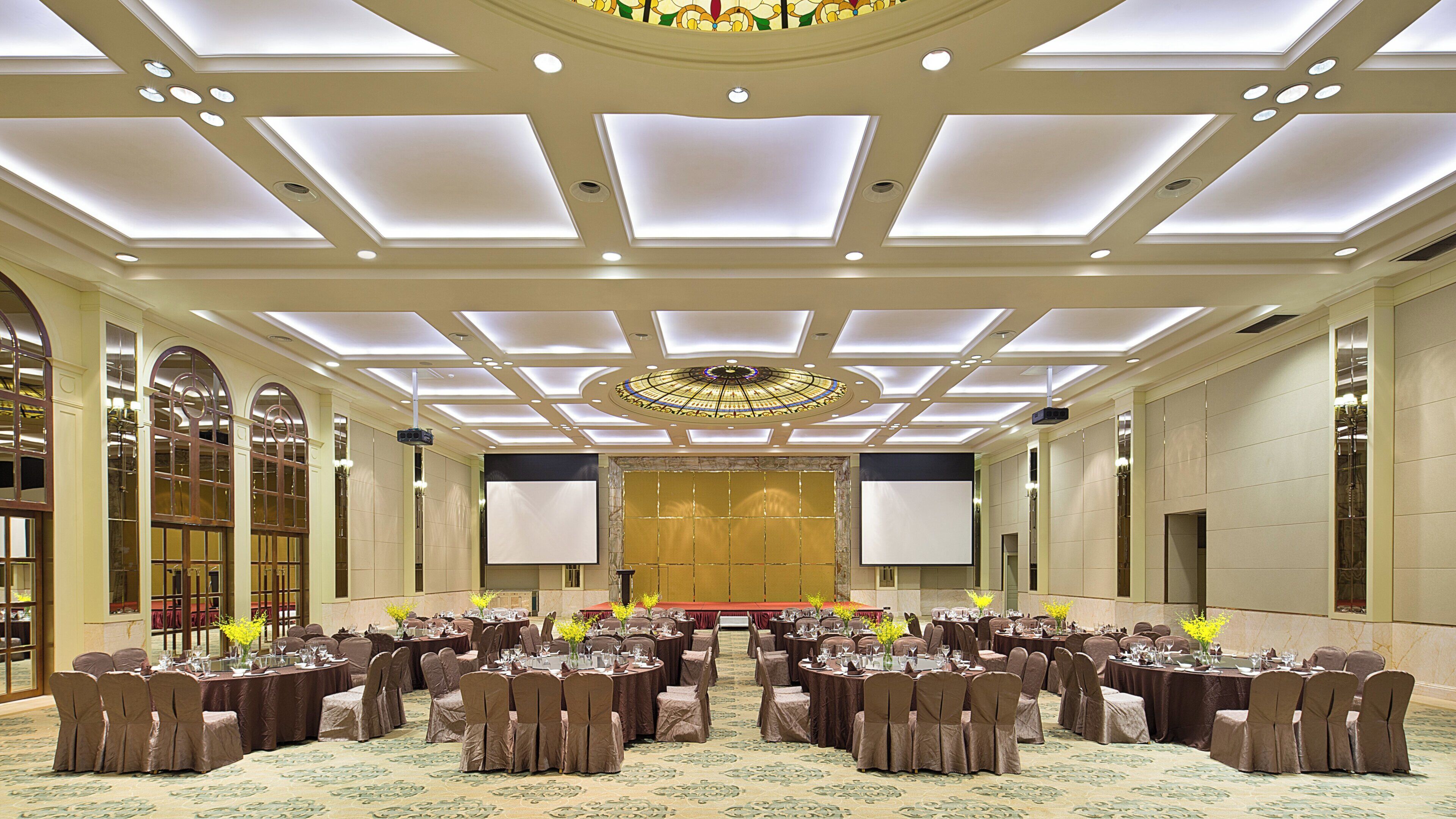 A banquet hall with round tables, chairs, and a stage with a large screen.