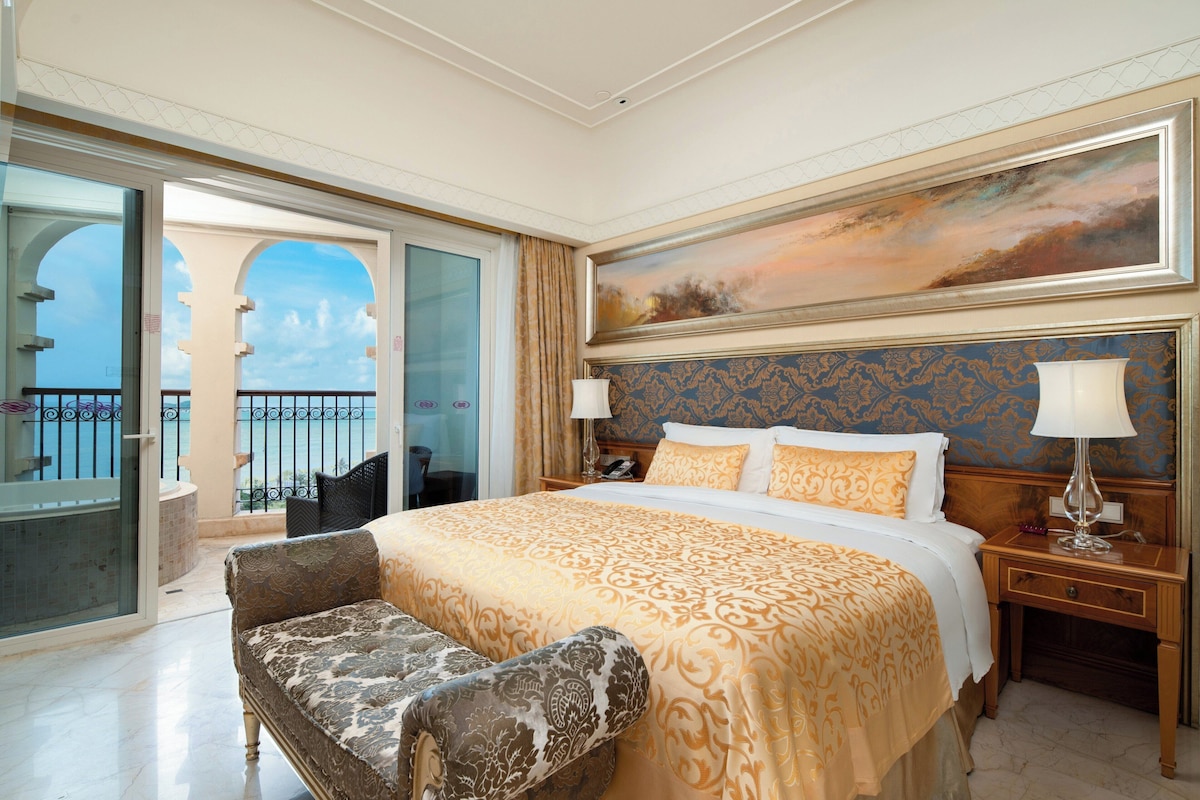 A hotel room with a large bed, a chair, a painting, and a balcony with a view.