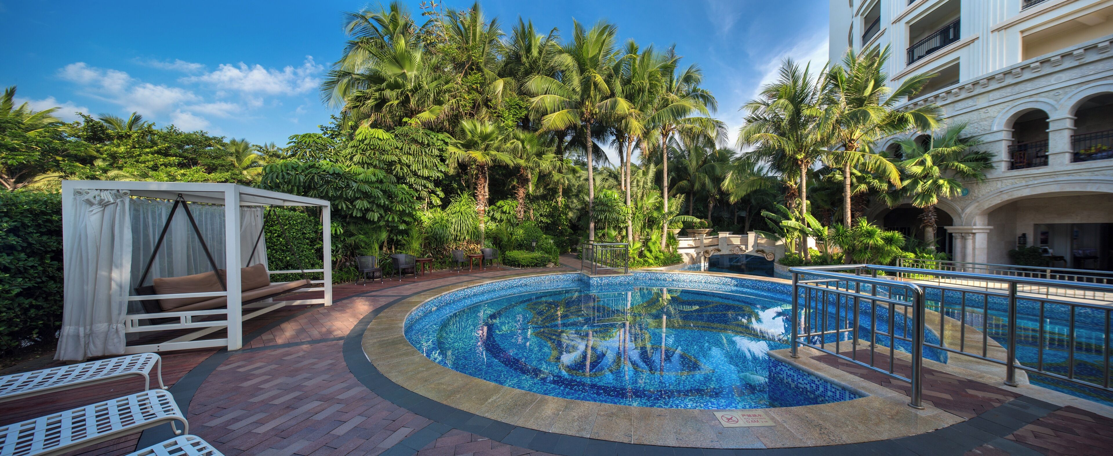 A pool area with a circular design, surrounded by palm trees and a white gazebo.