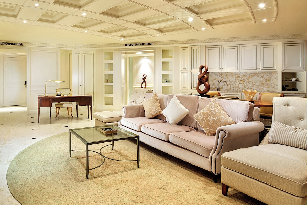 A spacious living room with a beige sofa, a glass coffee table, and a dining area with a marble backsplash.