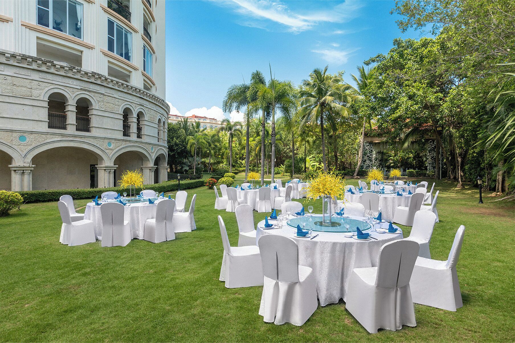 Un banquet en plein air avec des tables rondes, des chaises blanches et des fleurs jaunes.