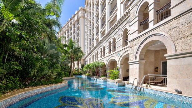 Une vaste piscine intérieure à l’eau d’un bleu limpide, entourée d’une végétation luxuriante et bordée d’un bâtiment classique dont la façade est ornée d’arcades et de balcons.