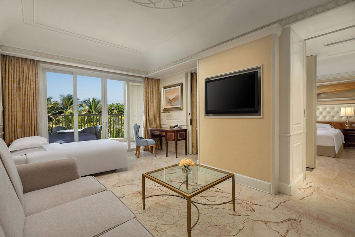 A hotel room with a bed, a sofa, a desk, a TV, and a balcony with a view of palm trees.
