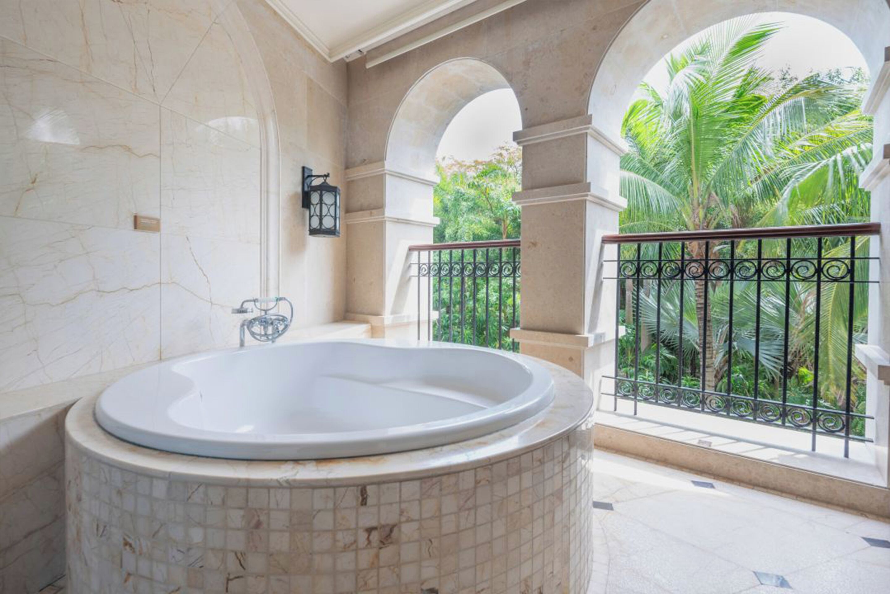 A bathroom with a large marble bathtub, a tiled floor, and a view of a balcony with a wrought iron railing and palm trees.