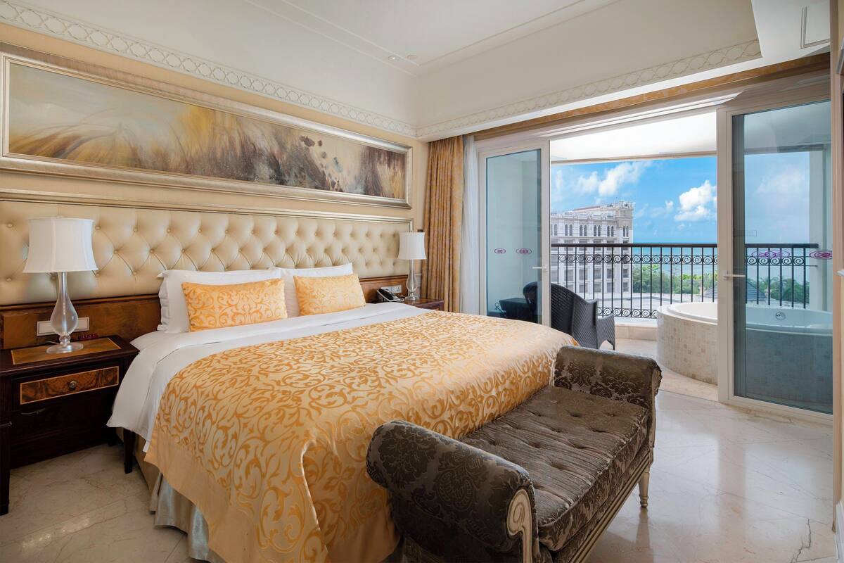 Une chambre d’hôtel avec un grand lit, une banquette, une table de chevet, une lampe et une vue sur un balcon doté d’une balustrade, avec un bâtiment en arrière-plan.