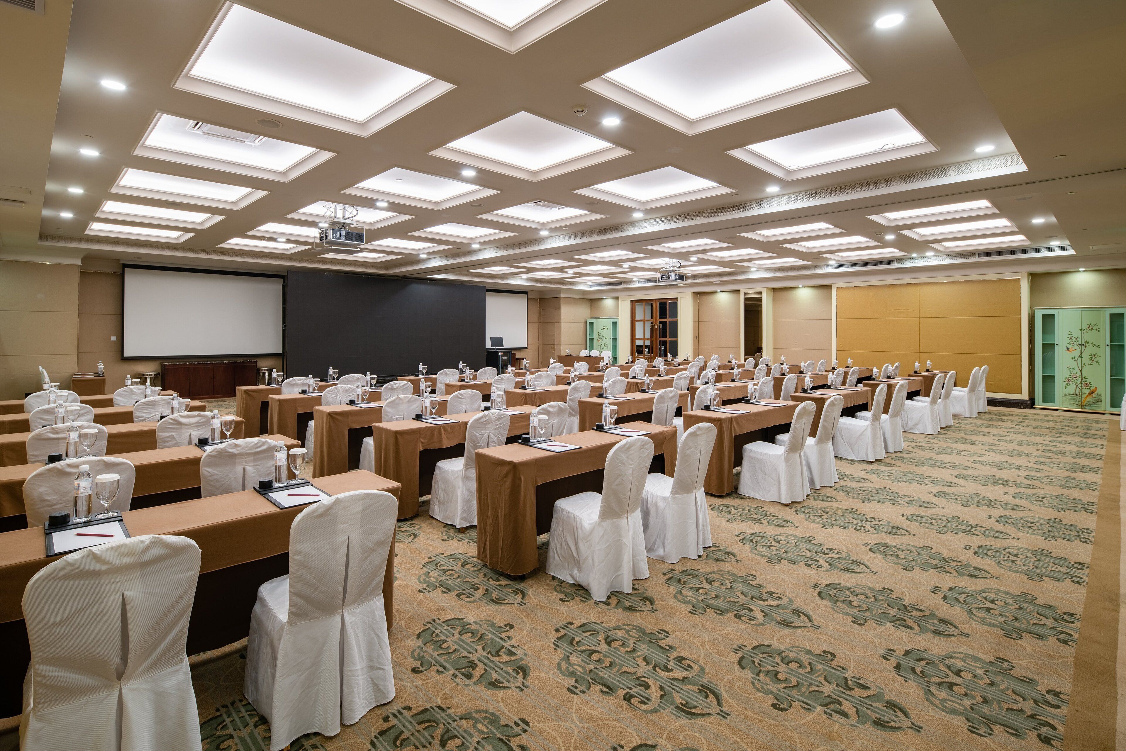 Un salón de conferencias con varias mesas y sillas, equipado con proyectores y pantallas, y piso alfombrado.