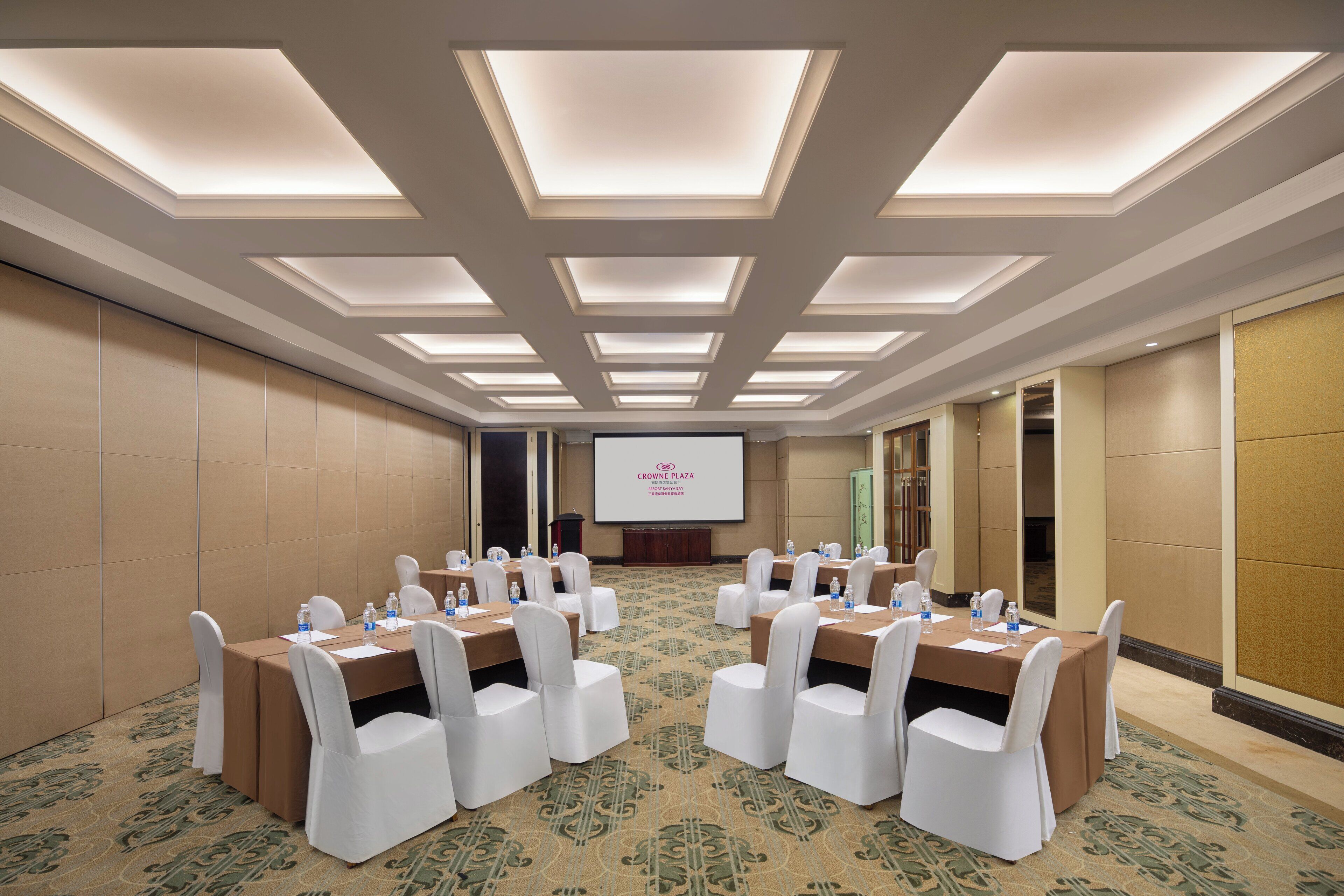 Une salle de conférence dotée d’un grand écran, de plusieurs tables avec des chaises et d’un tapis à motifs.