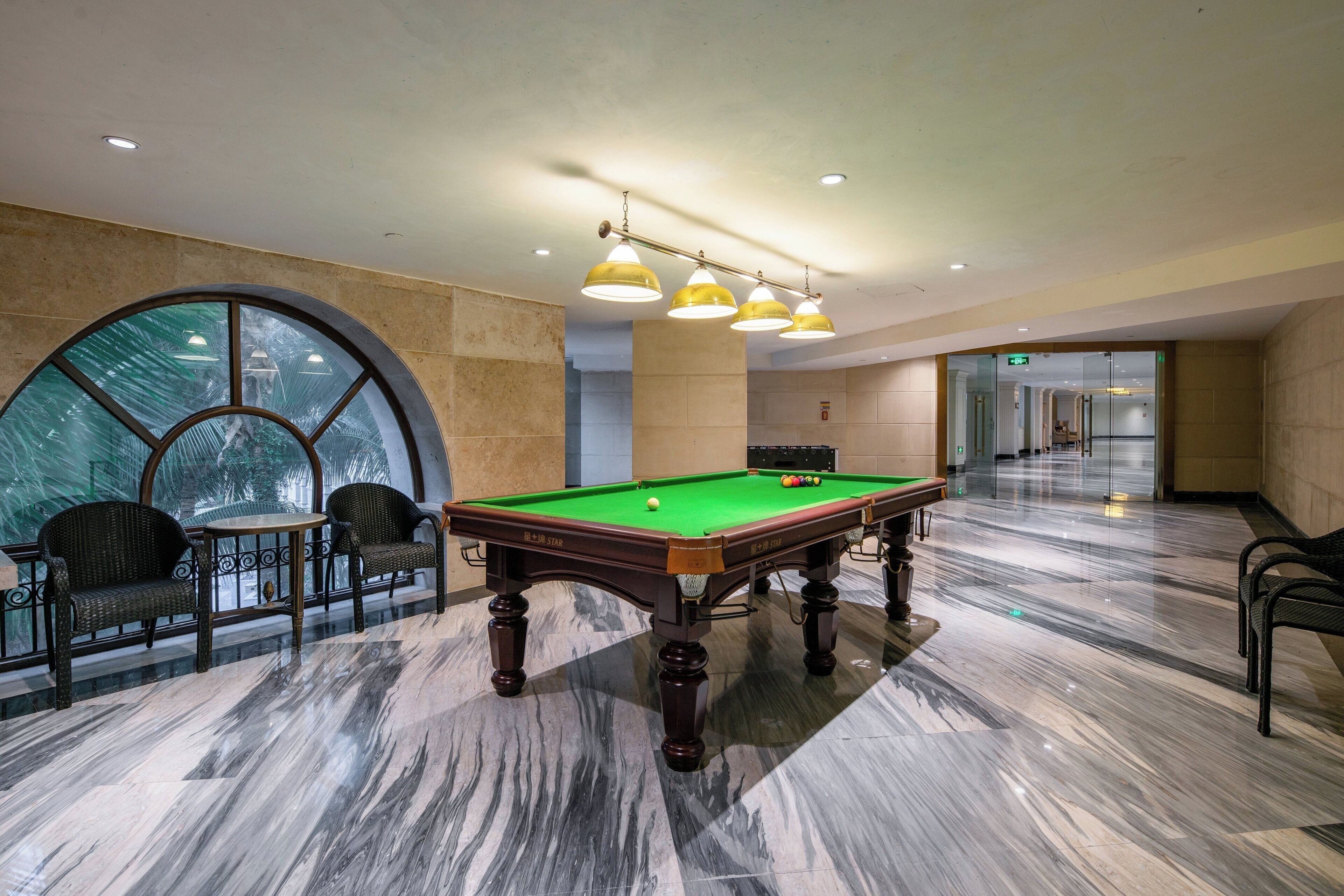 Une table de billard dans une pièce dotée d’un sol en marbre, d’une grande fenêtre et d’un coin salon avec des fauteuils.