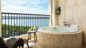 Un balcon avec vue sur la mer, un jacuzzi, des fauteuils en osier et une petite table agrémentée de fleurs et d’une boisson.