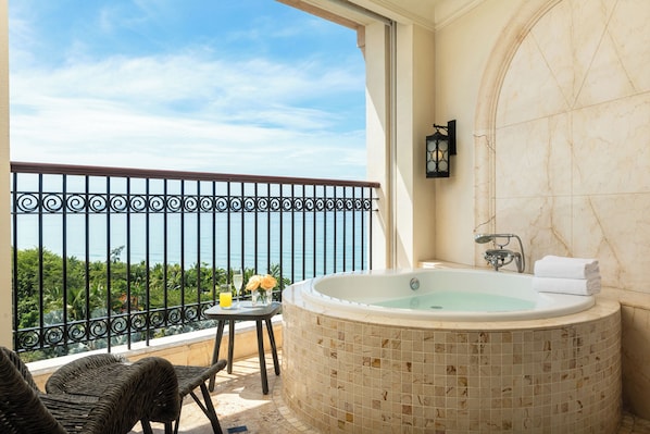 A balcony with a view of the sea, a hot tub, wicker chairs, and a small table with flowers and a drink.