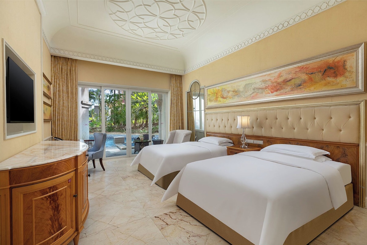 A hotel room with two beds, a desk, a chair, a TV, and a large window with a view of greenery.