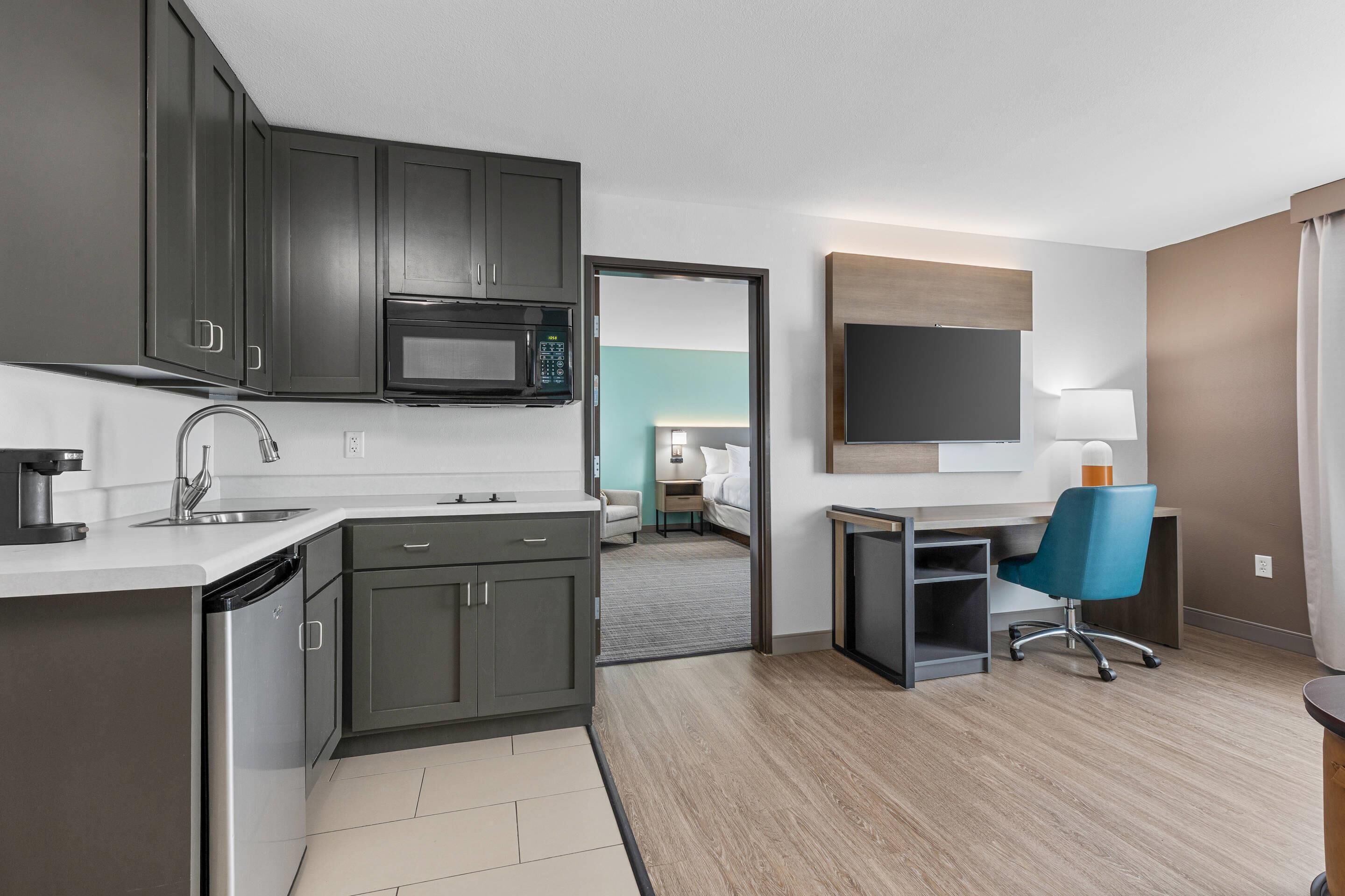 A modern hotel room with a kitchenette, a flat-screen TV, a desk with a chair, and a bed in the background.