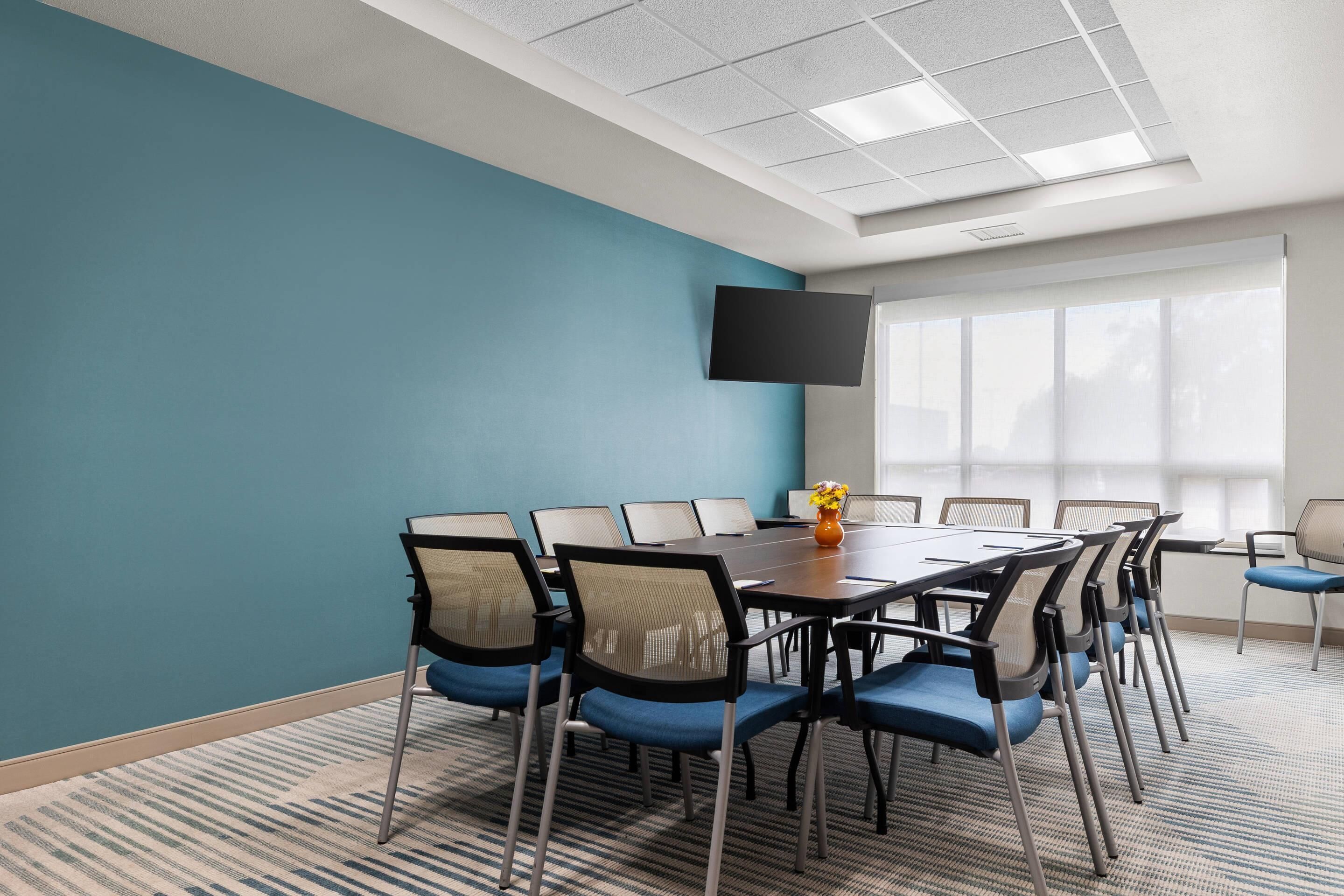 A conference room with a large table, chairs, a flat-screen TV, and a window with blinds.
