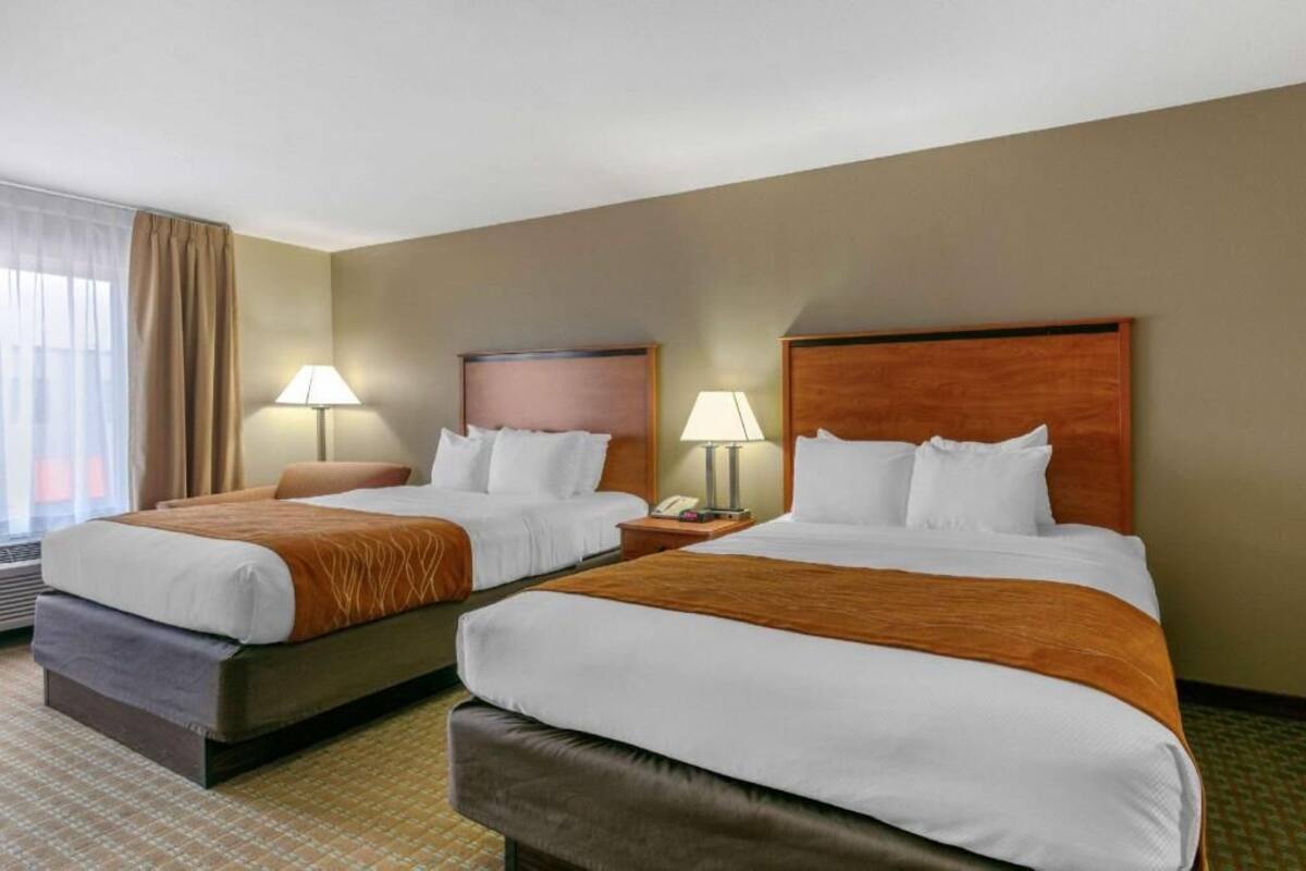 A hotel room with two beds, each with a wooden headboard, a bedside table with a lamp, and a window with curtains.