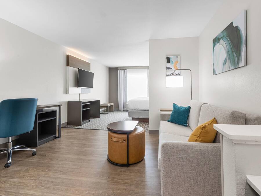 Una habitación de hotel moderna con televisión de pantalla plana, un sofá, un otoman redondos, un escritorio y una silla.