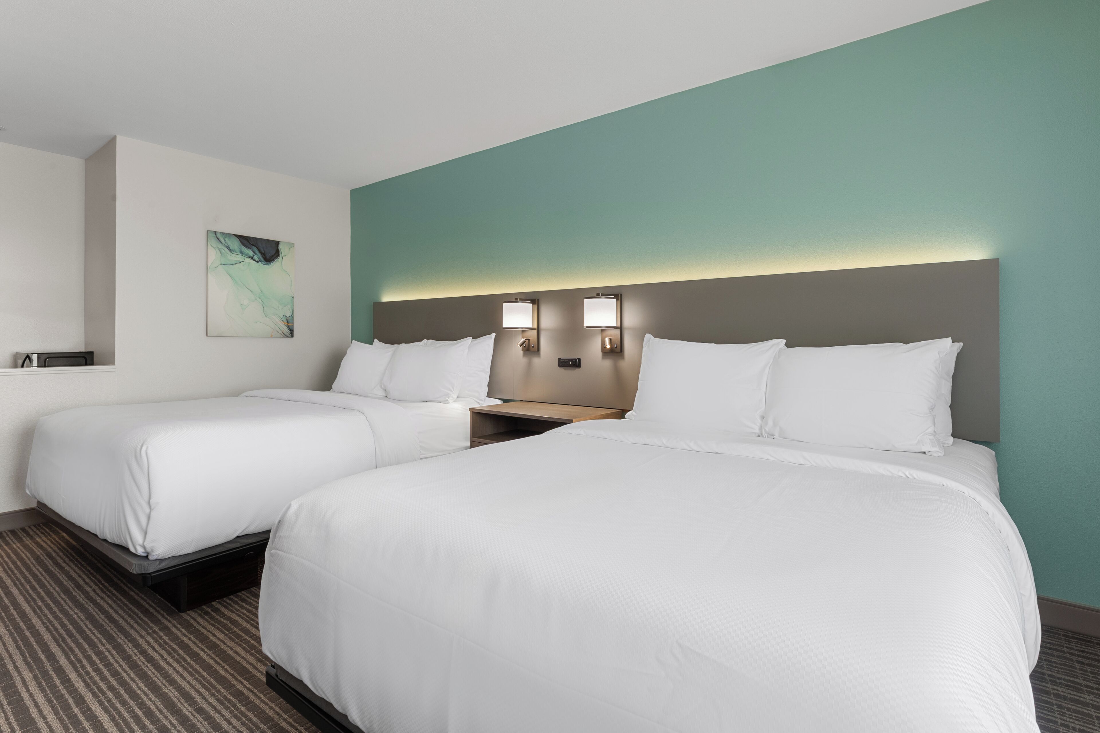 A hotel room with two beds, a nightstand, a wall-mounted lamp, and a painting.