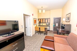 A compact living space with a flat-screen TV, a sofa, a dining table with chairs, a kitchen area, and a small desk.