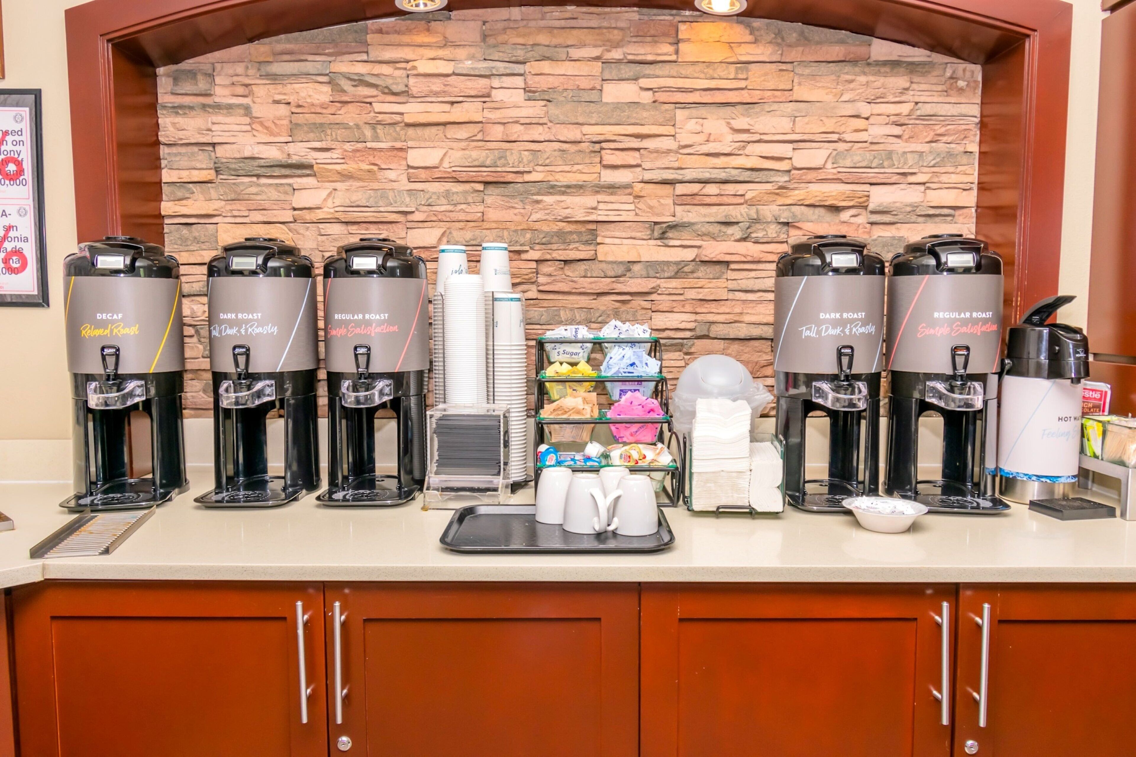 Una estación de café con varios dispensadores de café, un dispensador de azúcar y una bandeja con vasos desechables.