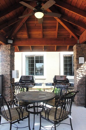 Un patio exterior con una mesa redonda y sillas, un ventilador de techo y vistas a una casa con grandes ventanales.