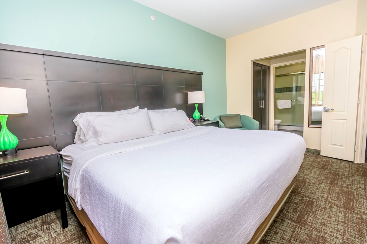A hotel room with a large bed, a nightstand with a lamp, a chair, and a bathroom visible through an open door.