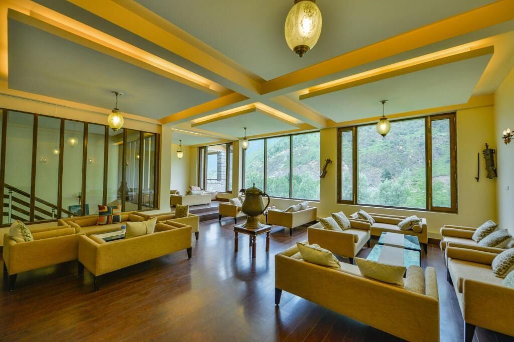 A spacious living area with multiple sofas and a coffee table, large windows offering a view of greenery, and a staircase leading to an upper level.