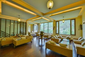 A spacious living area with multiple sofas and a coffee table, large windows offering a view of greenery, and a staircase leading to an upper level. - Heevan Villas Sonamarg (Gund)