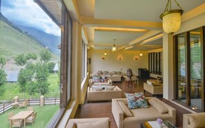 A spacious living area with multiple sofas and a television, large windows offering a view of a green landscape, and a balcony with outdoor seating. - Heevan Villas Sonamarg (Gund)